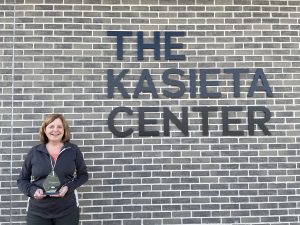 Karen holds BPNN's Champions in Action Award in front of a brick wall with metal lettering that says "The Kasieta Center."